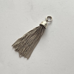 May include: A silver-toned metal tassel pendant featuring a smooth, rounded cap and a circular jump ring at the top. The tassel is composed of numerous fine, flexible chain strands that hang downward in a dense, uniform, and fan-like arrangement.
