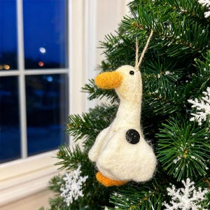 May include: A handmade, off-white goose ornament with an orange beak and feet, and a black button detail. The goose is hanging on a Christmas tree with green needles and artificial snow. A window is visible in the background.