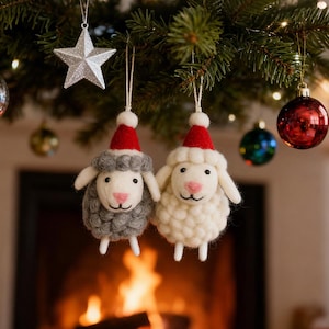 Op de afbeelding: Twee kerstversieringen in de vorm van schapen, één grijs en één wit, elk met een rode kerstmuts. Ze hangen aan een dennentak met andere ballen en een zilveren ster. De schapen hebben roze neuzen en zijn van wol.