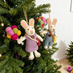 May include: Two felt bunny ornaments hanging on a Christmas tree. One bunny wears a purple sweater and holds a bunch of colorful felt ball balloons. The other bunny wears blue overalls and carries a yellow bag, also holding felt ball balloons.