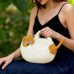 Könnte beinhalten: Weiß-orangefarbene Filz-Handtasche in Katzenform mit Henkel. Die Tasche hat ein Katzengesicht mit blauen Augen und einem schwarzen Kreuz als Mund. Sie hat einen orangenen Schwanz und einen kleinen orangenen Fleck an der Seite.