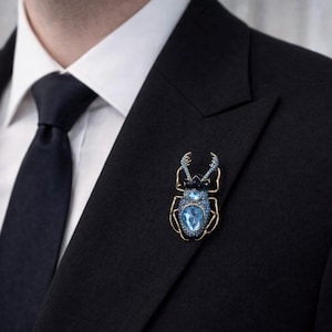 May include: A gold-toned beetle brooch with blue crystal accents, pinned to a black suit jacket. The brooch features a large, light blue crystal at the center of the body and smaller crystals outlining the wings and head.