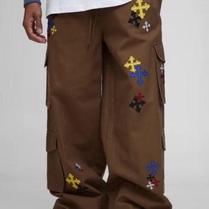 May include: Brown cargo pants with multiple pockets and colorful cross embroidery. The crosses are in yellow, blue, red, and black. The pants have a relaxed fit and a drawstring waist.