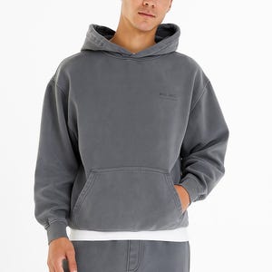 Puede incluir: Una sudadera con capucha gris con bolsillo de canguro y capucha con cordón. La palabra "WAGLUND" está impresa en el pecho. La persona lleva debajo una camiseta blanca y un pantalón de chándal gris a juego.