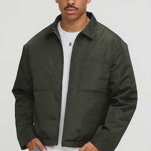 Puede incluir: Chaqueta verde oliva con cierre de cremallera y dos bolsillos delanteros. El modelo lleva una gorra negra con la palabra "Hen" en escritura blanca. El modelo también lleva una camiseta blanca y vaqueros azules.