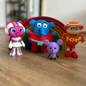 Puede incluir: Colección de figuras de personajes de dibujos animados coloridos, incluyendo un gato rosa, un elefante azul, un ratón morado y un mono naranja. Las figuras están dispuestas frente a un arcoíris de madera.