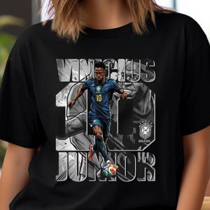 Könnte beinhalten: Schwarzes T-Shirt mit einer Grafik eines Fußballspielers in blauer Uniform, mit dem Namen "VINICIUS JR" und der Nummer "10" in Weiß. Die brasilianische Flagge und das Wort "BRASIL" sind ebenfalls zu sehen.