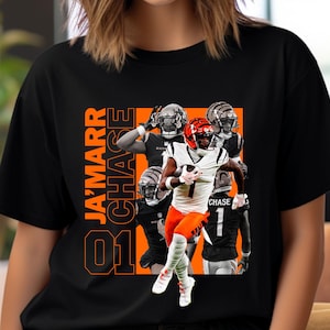 May include: Black t-shirt featuring a graphic of a football player in a white and orange uniform, with the name "Ja'Marr Chase" in orange block letters. The player is running with the ball, surrounded by other players.