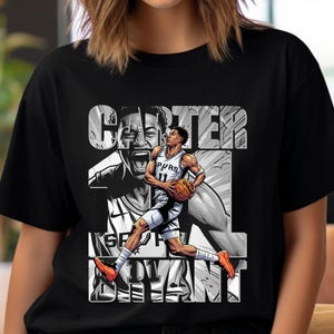 May include: Black t-shirt featuring a graphic of a basketball player in action, holding a basketball. The player is wearing a white and gray uniform with the text "SPURS" visible. The name "CARTER" is printed above the image.