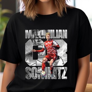 Könnte beinhalten: Ein schwarzes T-Shirt mit kurzen Ärmeln und einem Grafikdruck eines Fußballspielers in rot-weißem Trikot, der einen Ball kickt. Der Text MAXIMILIAN SOMNITZ ist in großen weißen Buchstaben über einem graustufigen Porträt des Spielers abgebildet.