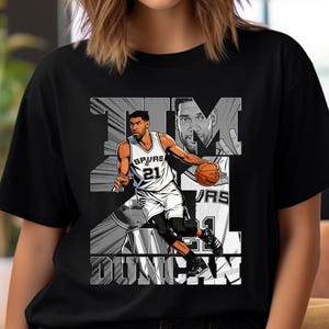 Peut inclure: T-shirt noir avec un graphisme en noir et blanc d'un joueur de basket-ball en uniforme des Spurs, avec le nom "TIM DUNCAN". Le design comprend un portrait du joueur. Style graphique et audacieux.