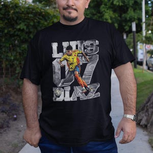 May include: Black t-shirt featuring a graphic of a soccer player in a yellow and orange uniform, with the name "Luis Diaz" and the number "7" in a stylized design. The player is depicted kicking a soccer ball.