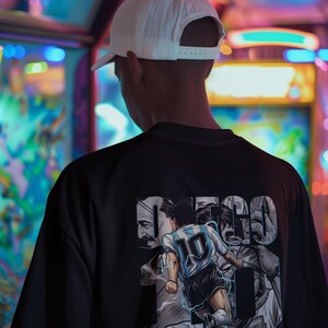 May include: Black t-shirt featuring a graphic of a soccer player in a blue and white jersey with the number 10. The shirt also has the name "MARADONA" and other text. A white baseball cap is worn.