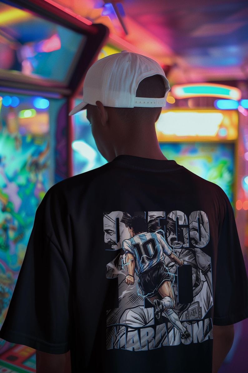 May include: Black t-shirt featuring a graphic of a soccer player in a blue and white jersey with the number 10. The shirt also has the name "MARADONA" and other text. A white baseball cap is worn.