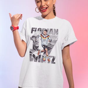 May include: White t-shirt featuring a graphic of a soccer player in a German national team uniform, with the name "FLORIAN" and the number "17" printed on it. The design includes a portrait and the name "WIRTZ".