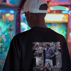 May include: Black t-shirt featuring a graphic of a soccer player in a blue and white uniform, with the number 10. The graphic includes the name ZIDANE. The person in the image is wearing a white baseball cap.