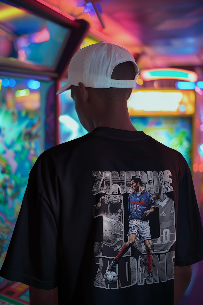 May include: Black t-shirt featuring a graphic of a soccer player in a blue and white uniform, with the number 10. The graphic includes the name ZIDANE. The person in the image is wearing a white baseball cap.