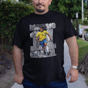 May include: Black t-shirt featuring a graphic of a soccer player in a yellow and blue uniform, with the name "ROMARIO" and the word "BRAZIL" in a gray, stylized font. The player is depicted in action, kicking a soccer ball.
