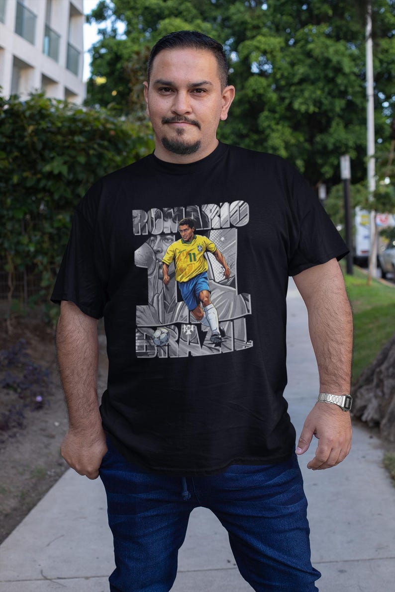 May include: Black t-shirt featuring a graphic of a soccer player in a yellow and blue uniform, with the name "ROMARIO" and the word "BRAZIL" in a gray, stylized font. The player is depicted in action, kicking a soccer ball.