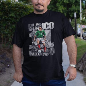 May include: Black t-shirt featuring a graphic of a soccer player in a green and red uniform, with the text "BLANCO 10 MEXICO 98". The player is depicted kicking a soccer ball. The shirt is worn by a person.