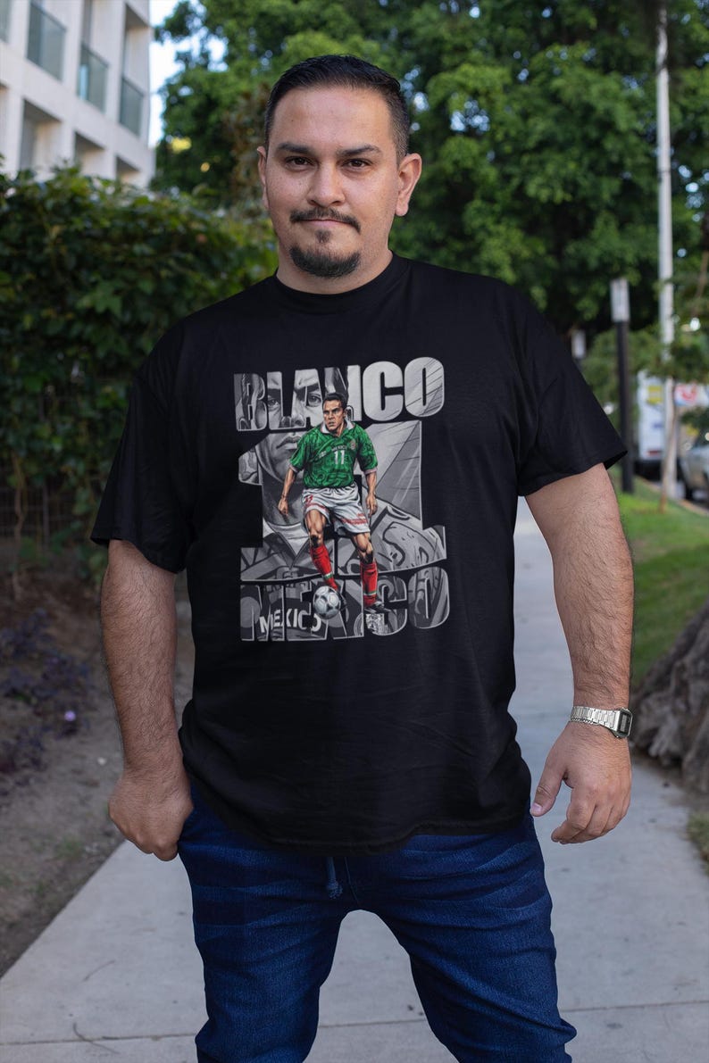 May include: Black t-shirt featuring a graphic of a soccer player in a green and red uniform, with the text "BLANCO 10 MEXICO 98". The player is depicted kicking a soccer ball. The shirt is worn by a person.