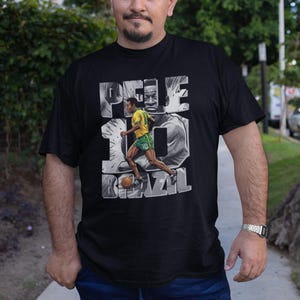 May include: Black t-shirt featuring a graphic of Pel&eacute; in a yellow and green soccer uniform, with the number 10 and the word BRAZIL. The design includes a grayscale image of Pel&eacute;'s face.