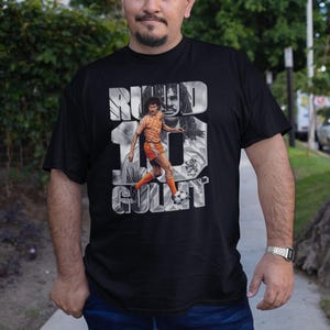 May include: Black t-shirt featuring a graphic of Ruud Gullit in an orange soccer uniform, with the text "RUUD 10 GULLIT" in a stylized font. The shirt is worn by a person.