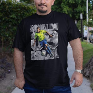 May include: Black t-shirt featuring a graphic of a soccer player in a yellow and blue uniform kicking a ball. The shirt has the text "RONALDO 9 BRAZIL" in a stylized font.