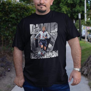 May include: Black t-shirt featuring a graphic of David Beckham in an England soccer uniform. The design includes the name "DAVID BECKHAM" and the number 7. The shirt is worn by a person in blue jeans.