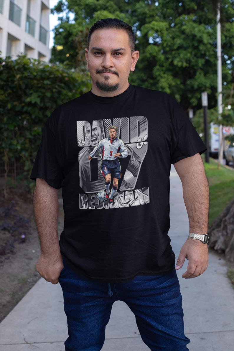 May include: Black t-shirt featuring a graphic of David Beckham in an England soccer uniform. The design includes the name "DAVID BECKHAM" and the number 7. The shirt is worn by a person in blue jeans.