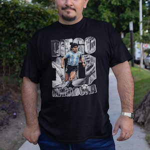 May include: Black t-shirt featuring a grayscale image of Diego Maradona in action, with the name "DIEGO MARADONA" printed above. The shirt is worn by a person in blue jeans.