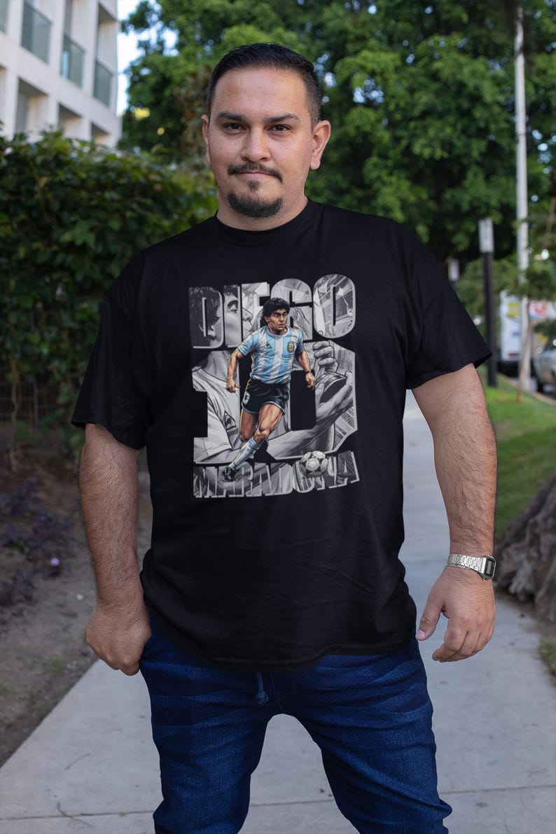 May include: Black t-shirt featuring a grayscale image of Diego Maradona in action, with the name "DIEGO MARADONA" printed above. The shirt is worn by a person in blue jeans.