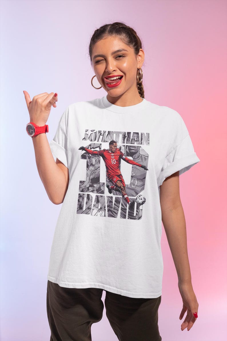 May include: White t-shirt featuring a graphic of a soccer player in a red uniform, with the name "JONATHAN DAVID" and the number 10. The design has a grayscale background with the player in action. The shirt is worn by a person.