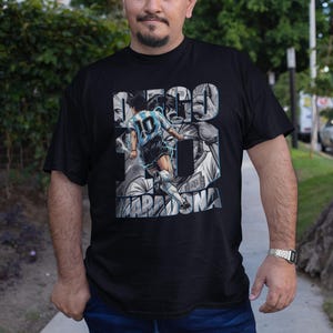 May include: Black t-shirt featuring a graphic of a soccer player in a light blue and white uniform, with the name "MARADONA" and the number 10. The shirt has a portrait design and the name "DIEGO" in large letters.