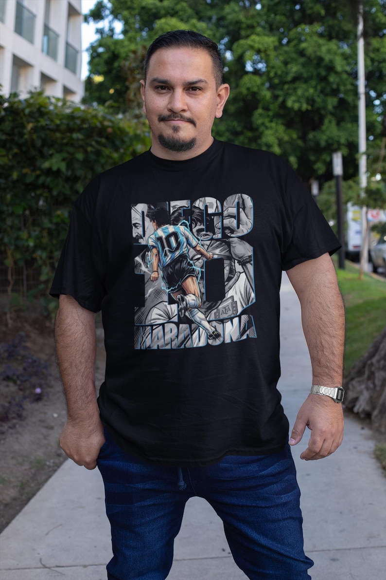 May include: Black t-shirt featuring a graphic of a soccer player in a light blue and white uniform, with the name "MARADONA" and the number 10. The shirt has a portrait design and the name "DIEGO" in large letters.