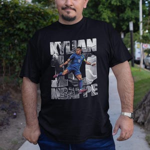 May include: Black t-shirt featuring a graphic of Kylian Mbapp&eacute; in a blue soccer uniform, with the name "KYLIAN" above and "MBAPPE" below. The number 10 is also visible. The shirt is worn by a person.