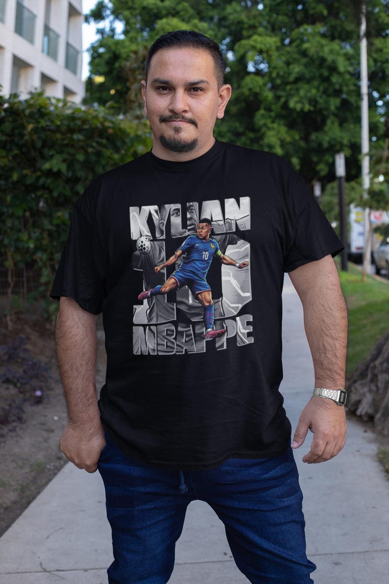 May include: Black t-shirt featuring a graphic of Kylian Mbapp&eacute; in a blue soccer uniform, with the name "KYLIAN" above and "MBAPPE" below. The number 10 is also visible. The shirt is worn by a person.