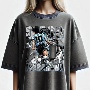 May include: Gray t-shirt featuring a graphic of a soccer player in action, wearing a blue and white jersey with the number 10. The design includes the name "DIEGO MARADONA" in a stylized font, with additional images of the player.