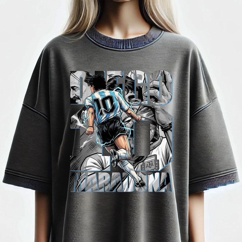 May include: Gray t-shirt featuring a graphic of a soccer player in action, wearing a blue and white jersey with the number 10. The design includes the name "DIEGO MARADONA" in a stylized font, with additional images of the player.