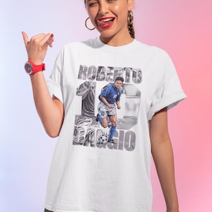 May include: White t-shirt featuring a graphic of Roberto Baggio in a blue soccer uniform, with the name "Roberto Baggio" and the number 10. The design includes a soccer ball and a stylized background.