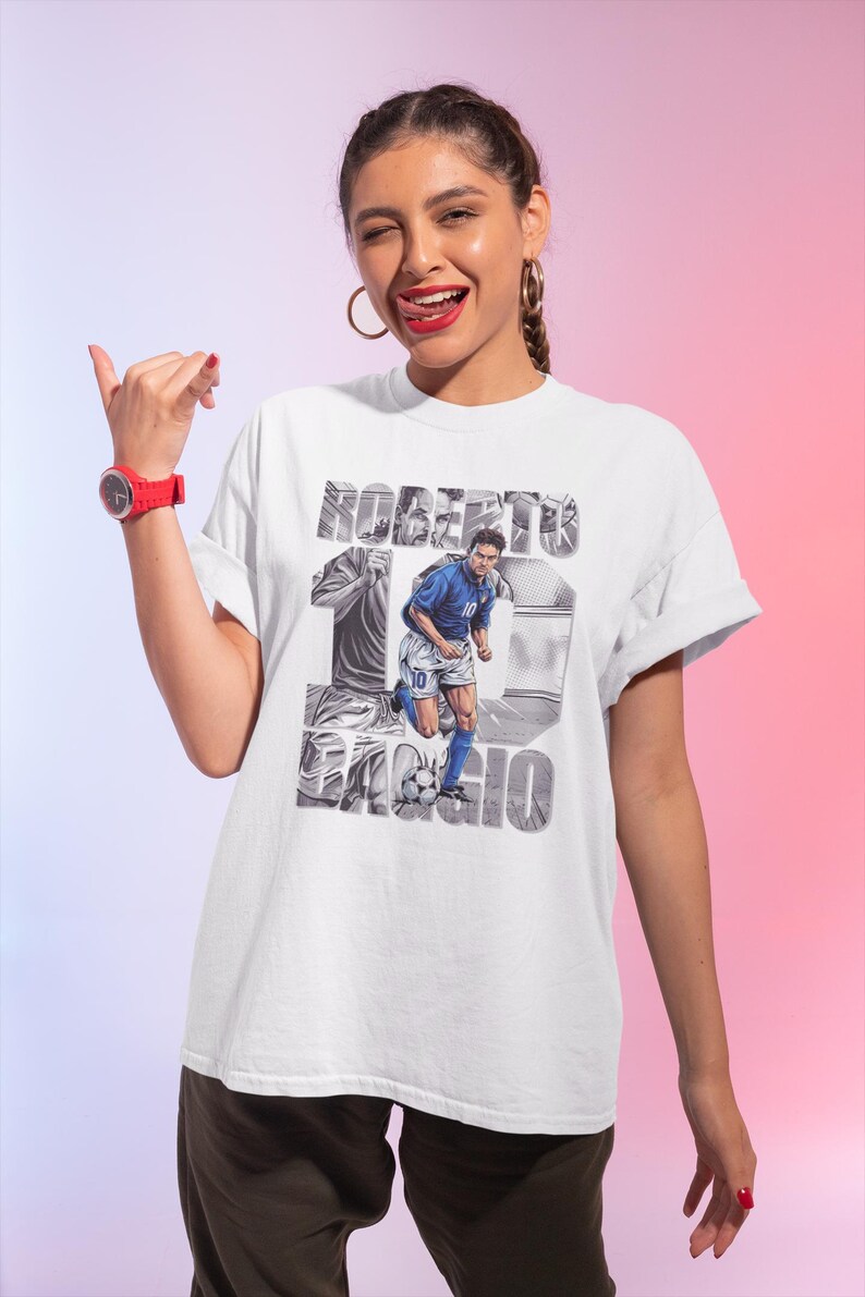 May include: White t-shirt featuring a graphic of Roberto Baggio in a blue soccer uniform, with the name "Roberto Baggio" and the number 10. The design includes a soccer ball and a stylized background.