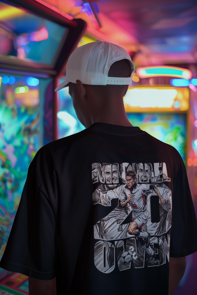 May include: Black t-shirt featuring a graphic of a soccer player with the name "OWEN" and the number 20. The design includes the text "ENGLAND FRANCE 1998". A white baseball cap is worn.