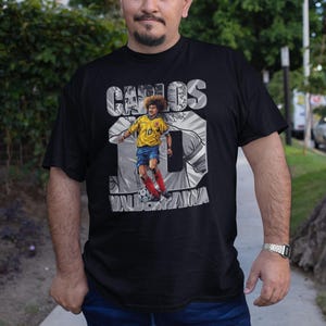 May include: Black t-shirt featuring a graphic of a soccer player in a yellow and blue uniform, with the name "CARLOS" above and the number "10" prominently displayed. The text "VALDERRAMA" is below the image. The shirt is worn by a person.