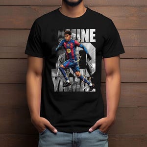 May include: Black t-shirt featuring a graphic of a soccer player in a blue and red uniform, with the name "LAMINE YAMAL" in white. The design includes the number 27 and a Spotify logo. The background is a grayscale image of a crowd.