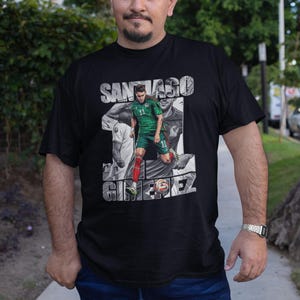 May include: Black t-shirt featuring a graphic of a soccer player in a green uniform with the name "SANTIAGO GIMENEZ" in white. The player is depicted in action, with a blurred background. The shirt is worn by a person.