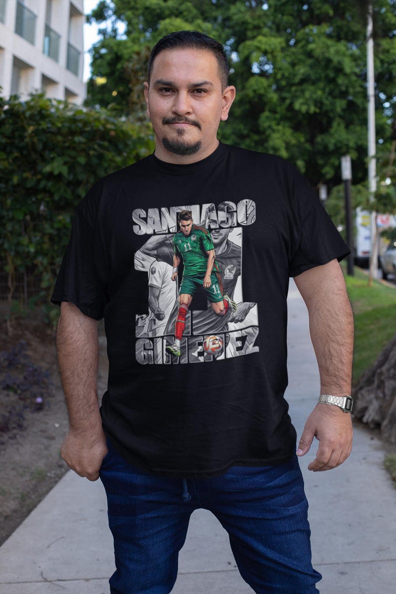May include: Black t-shirt featuring a graphic of a soccer player in a green uniform with the name "SANTIAGO GIMENEZ" in white. The player is depicted in action, with a blurred background. The shirt is worn by a person.