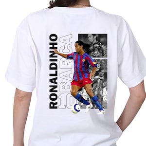 May include: White t-shirt featuring a color image of soccer player Ronaldinho in a Barcelona uniform, with the text "RONALDINHO BARCA 10" in black and white. Includes black and white images of Ronaldinho.