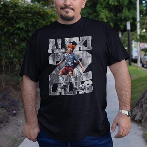 May include: Black t-shirt featuring a graphic of a soccer player with the name "ALEXI LALAS" and the number 22. The player is depicted in a dynamic pose, kicking a soccer ball. The shirt is worn by a person standing outdoors.