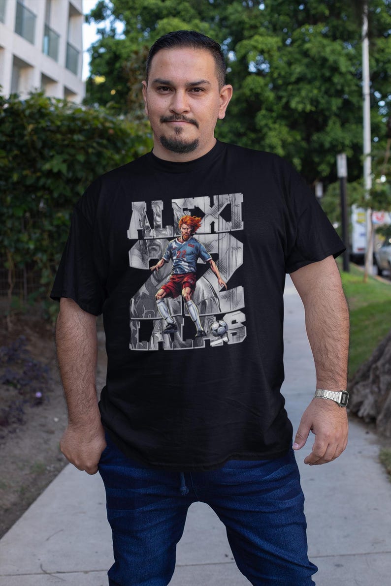 May include: Black t-shirt featuring a graphic of a soccer player with the name "ALEXI LALAS" and the number 22. The player is depicted in a dynamic pose, kicking a soccer ball. The shirt is worn by a person standing outdoors.