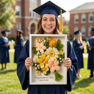 May include: A framed floral artwork with a white border, featuring a bouquet of colorful yarn flowers in shades of yellow, peach, and white. The text "Congratulations Kate Class Of 2026" is printed at the bottom. The person is wearing a graduation gown and cap.